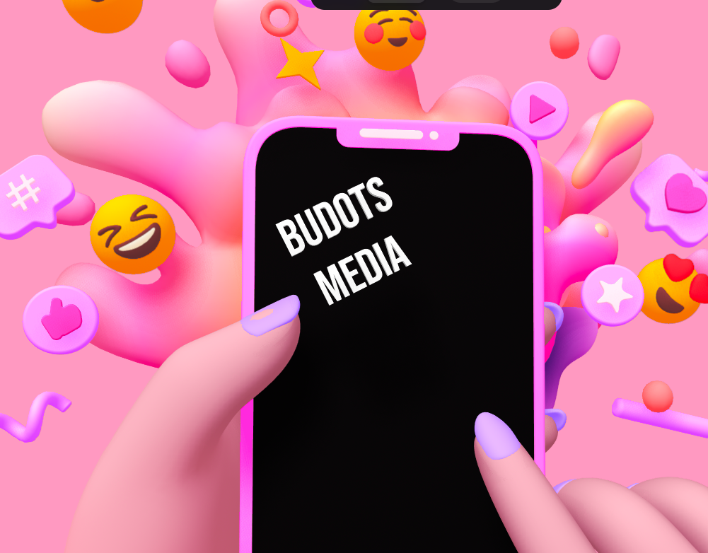 Illustration of a hand holding a smartphone with the screen displaying 'BUDOTS MEDIA'. The background is pink with various colorful 3D emojis and icons, such as laughing faces, hearts, thumbs up, and play buttons, floating around. The overall design is playful and vibrant, representing social media and digital content creation
